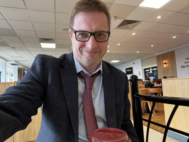 Photo of me sat in a claret and blue tweed, claret tie, blues shirt, and Burnley socks at knutsford services - nursing a cappuccino from Pret