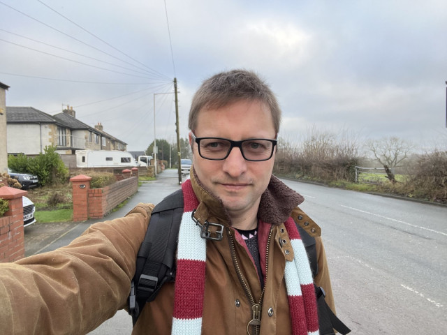 Me stood in Burnley wearing claret and blue scarf knitted by @craftedbypanda and Barbour Jacket.  Houses to the left, fields to the right with a road running through towards Pendle Hill under light grey sky.