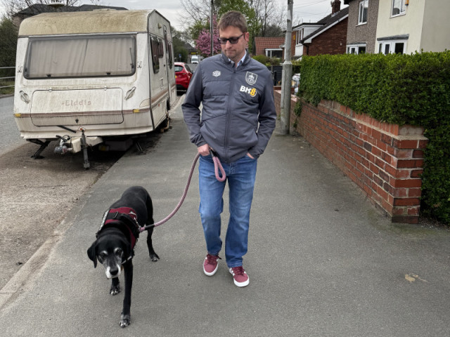 Old black pointer walks with me dressed in claret shoes, jeans, and a grey jacket, with a caravan covered in algae beyond
