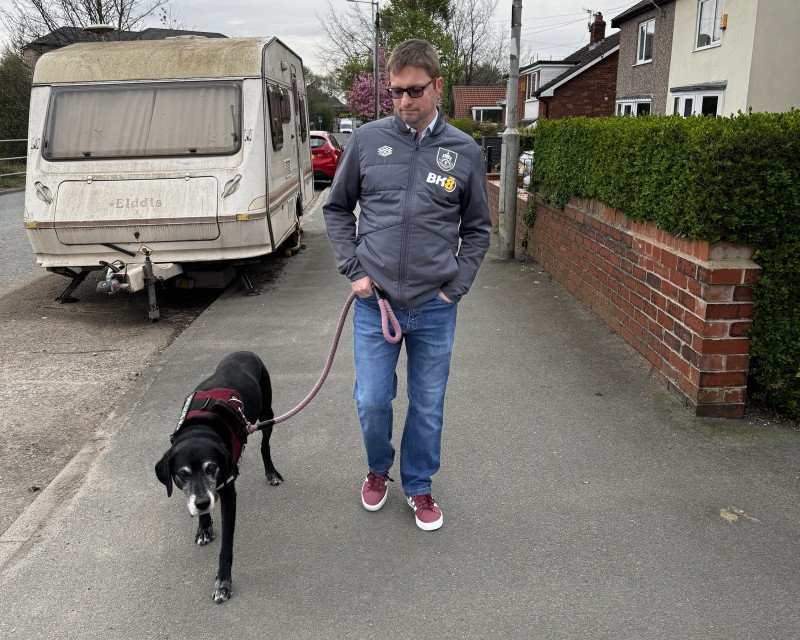 Old black pointer walks with me dressed in claret shoes, jeans, and a grey jacket, with a caravan covered in algae beyond