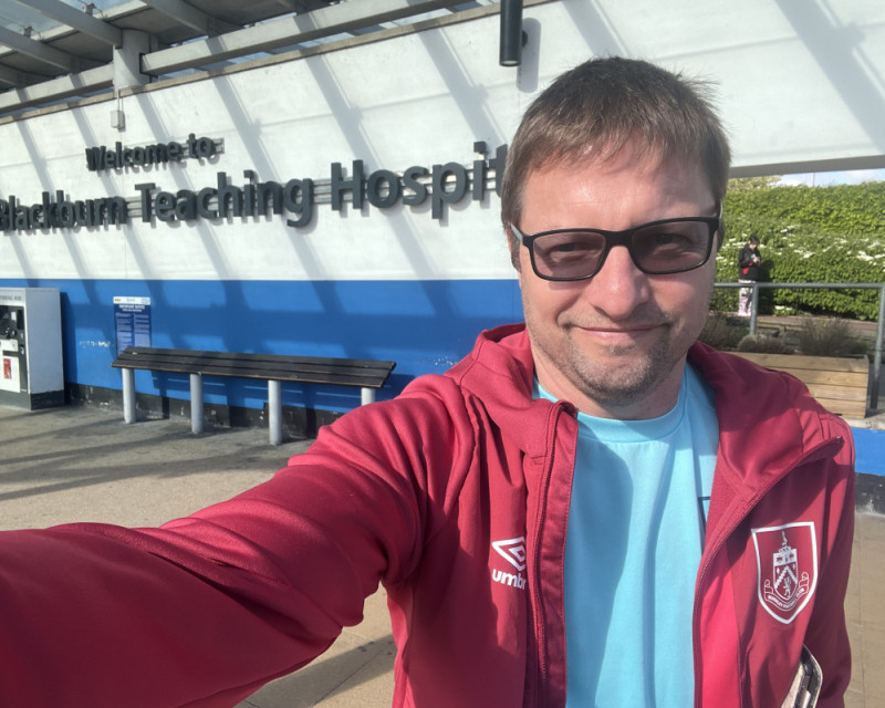 Me, stood in front of the Royal Bl@ckburn Hospital entry banner, in a branded claret Burnley soft shell and turquoise Burnley training top.